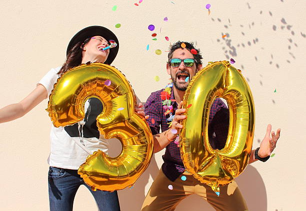 Cheerful couple celebrates a thirty years birthday Cheerful couple celebrates a thirty years birthday with big golden balloons and colorful little pieces of paper in the air. 30 34 years stock pictures, royalty-free photos & images