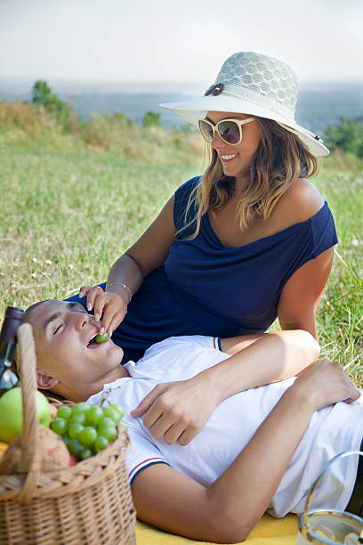 Woman feeding her boyfriend with grapes at picnic. Young couple enjoying at picnic in nature during beautiful summer day. Smiling woman is feeding her boyfriend with grapes while he's lying down in her lap. feeding grapes stock pictures, royalty-free photos & images
