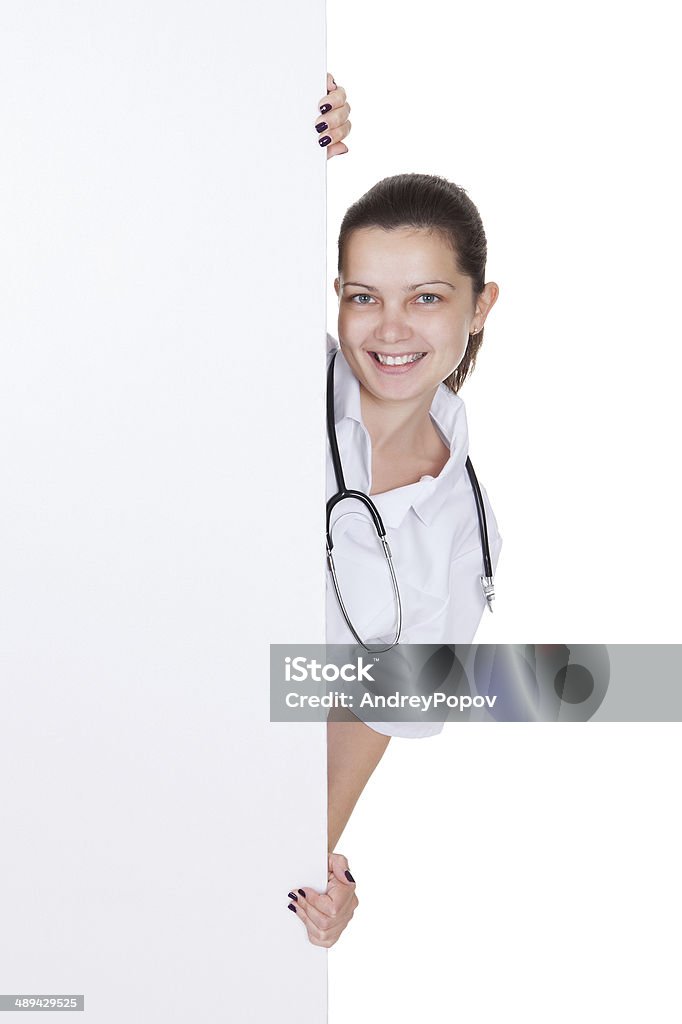 Doctor or nurse with blank signboard Attractive charming young female doctor or nurse with blank white signboard with copyspace peering around the side as she holds it up Adult Stock Photo