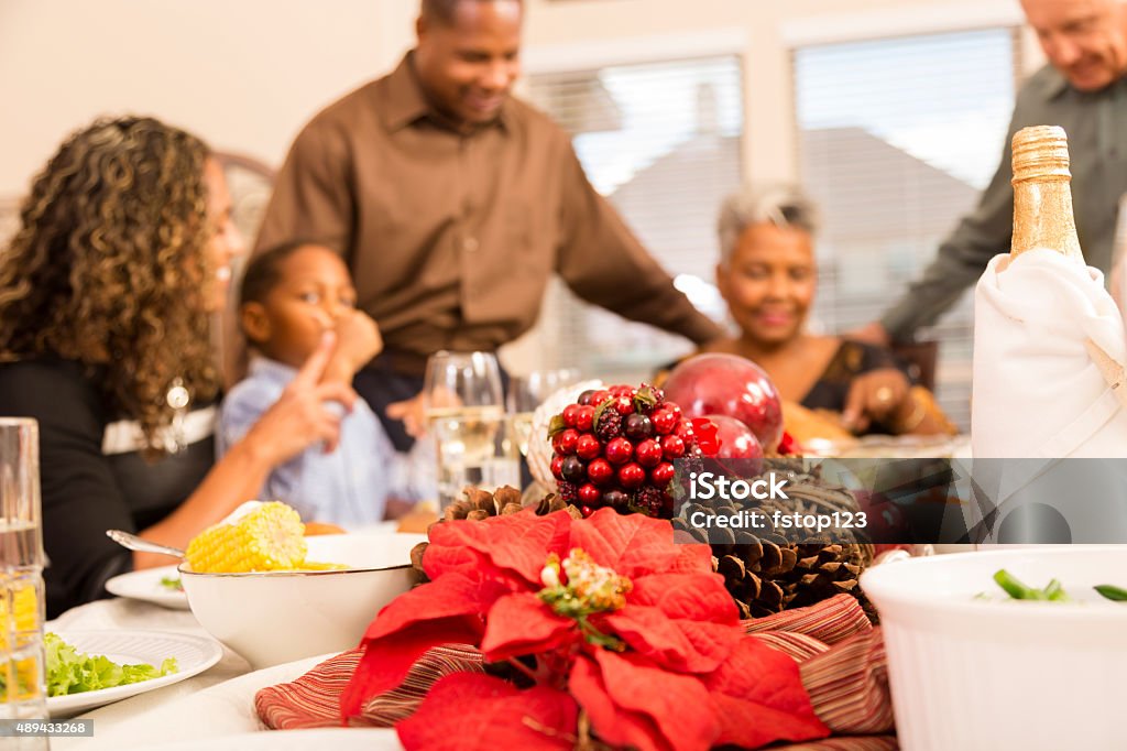 Family gather around the dinner table at Christmas time. Family gathers together for a holiday meal.  African descent little boy in mother's lap at dinner table while dad, grandmother and grandfather are in the background cutting the turkey.  Selective focus on Christmas poinsettia centerpice on table. Poinsettia Stock Photo