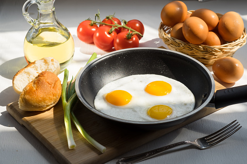 Three scrambled eggs on a frying pan with green onion, fork, bread, a basket eggs, tomatoes, and a bottle of olive oil.
