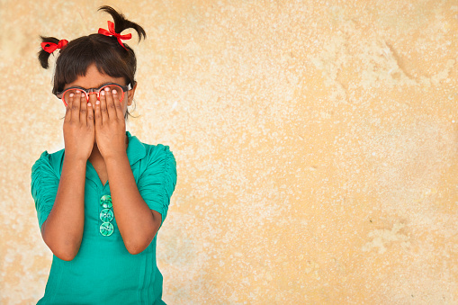 Cute, elementary age little girl wearing glasses and pigtails with hands covering her face in shame, guilt, embarrasment, nervousness, surprise, fear, or playing hide and seek.  The girl is of Latin, Indian or Asian descent.  Beige background.  Copyspace. 