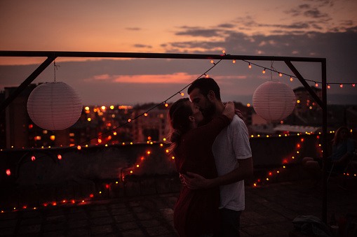 Young couple is kissing on the rooftop of the building, and sharing their love and affection under the evening sky 