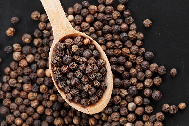 Black pepper whole in wooden spoon on black stone table Closeup black pepper whole in wooden spoon on black stone table, top view acrid taste stock pictures, royalty-free photos & images