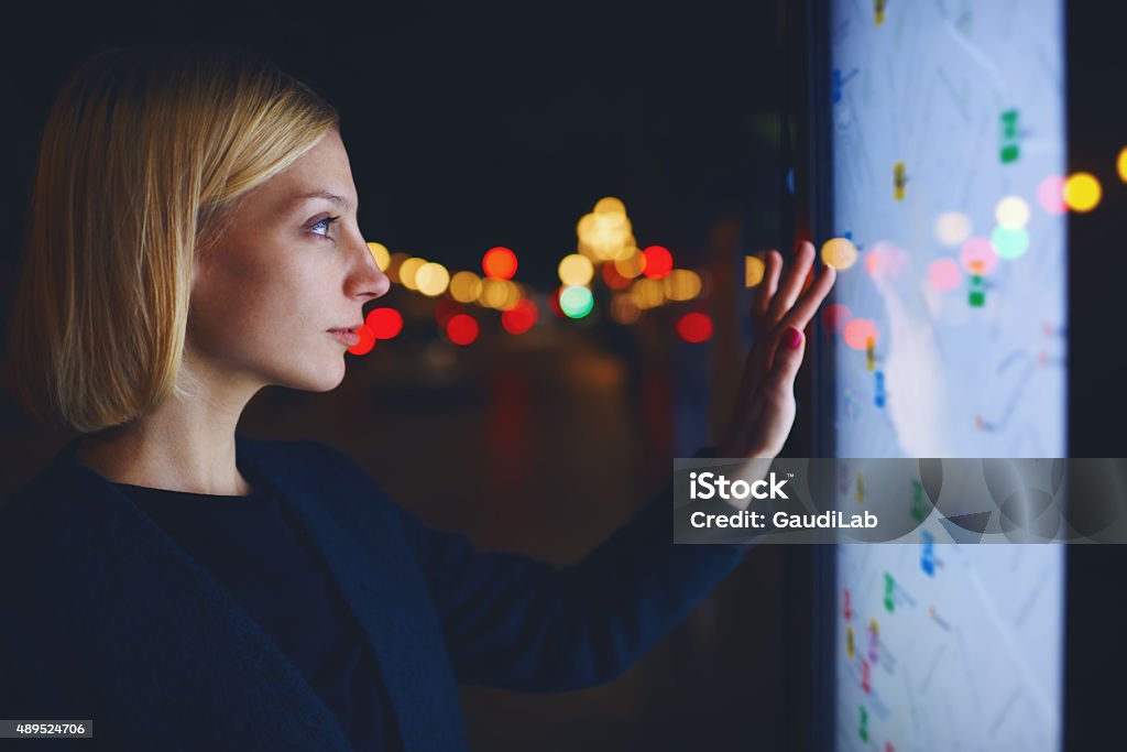 Charming female standing front big digital screen with city map Young female tourist using smart city gadget to get direction in Barcelona central, female in night city standing front big digital screen with city map routes and locations shown on it,filtered image Touch Screen Stock Photo
