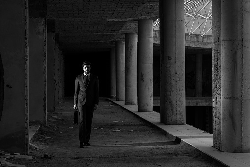 Full length black and white portrait of lonely businessman wearing suit in abandoned modern building.Group of columns are seen in diminishing perspective.Model is standing on the left side of horizontal frame.Main light is positioned on the left side of model.Shot with a full frame DSLR camera.