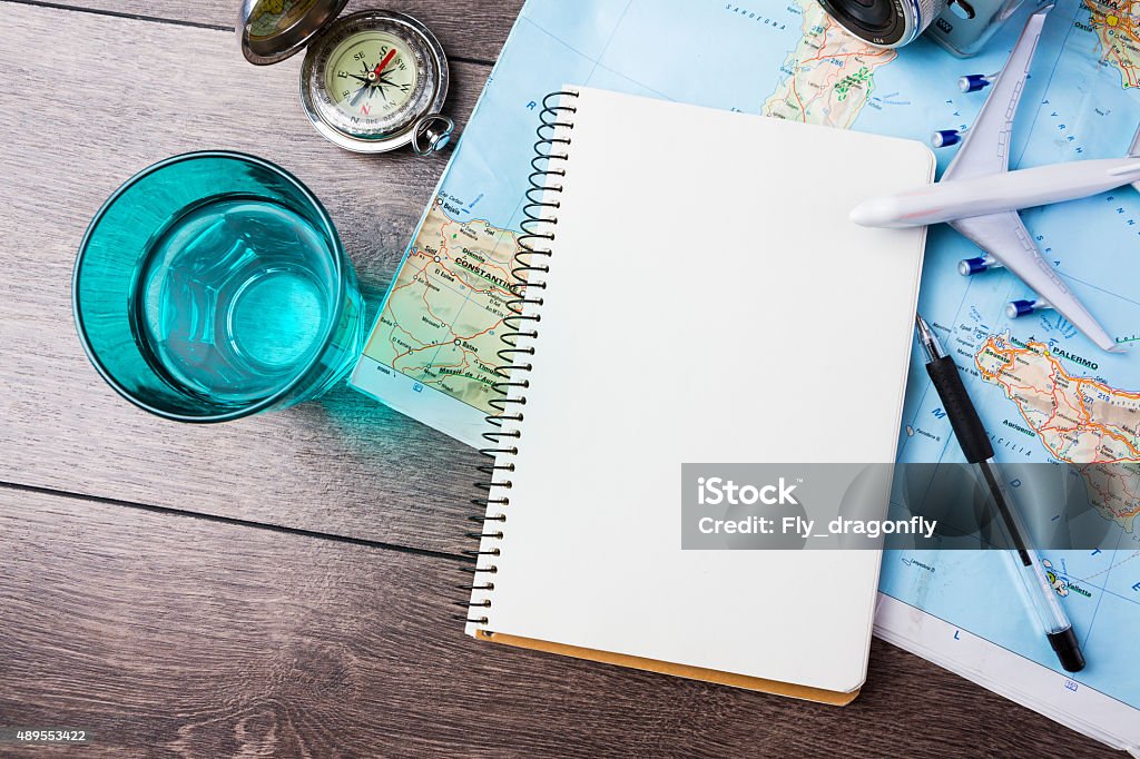 Travel theme tools travel , trip vacation, tourism mockup - close up of compass, glass of water note pad, pen and toy airplane and touristic map on wooden table. Empty space you can place your text or information. Travel Stock Photo