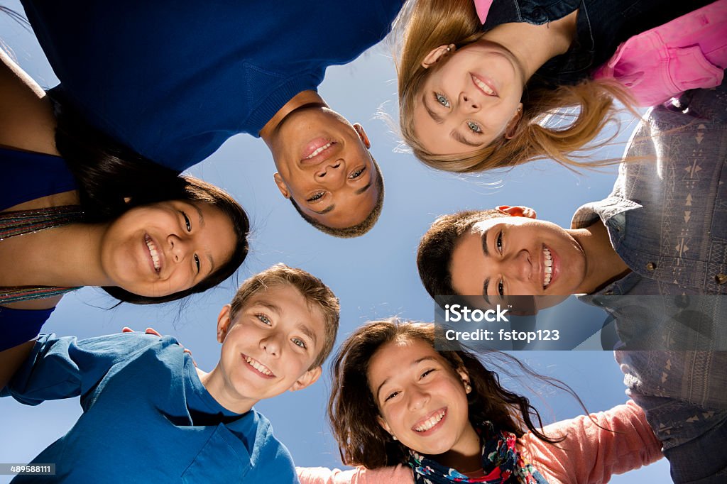 Teenagers: Diverse group of friends huddle outside together. Blue sky. Multi-ethnic group of teenage friends hang out, huddle together with arms around each other outdoors. Blue sky background.  Junior High Student Stock Photo