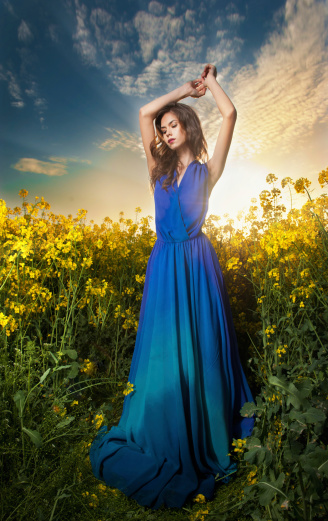 Fashion beautiful young woman in blue dress posing outdoor with cloudy dramatic sky in background. Attractive long hair brunette girl with elegant dress posing in canola field during sunset.
