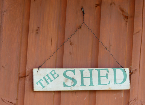 Photo showing homemade stenciled sign on door of wooden garden shed, simply saying 'The Shed', hanging on a piece of green garden wire.