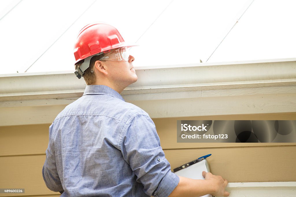 Inspector or blue collar worker examines building roof.  Outdoors. Repairman, building inspector, exterminator, engineer, insurance adjuster, or other blue collar worker has just begun his examination of a building/home's exterior walls, roof, and foundation. He wears a red hard hat and clear safety glasses and holds a his clipboard.   Rooftop Stock Photo