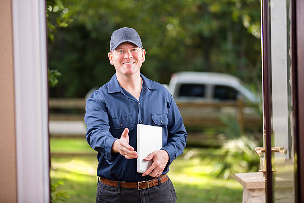 Service Industry: Repairman at customer's front door. Digital tablet. Handshake. Mid-adult repairman or blue collar/service industry worker makes service/house call at customer's front door. He holds his digital tablet with work invoices and reaches out to shake hands.  Inspector, exterminator, electrician. He wears a navy colored uniform. Service truck seen in background parked on road.  electrician smiling stock pictures, royalty-free photos & images