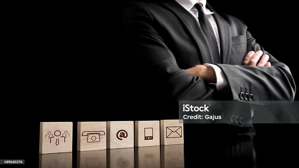 Businessman standing beside wooden blocks with drawings of commu Mid section of businessman wearing black suit, white shirt and black tie with arms crossed standing beside wooden blocks with drawings of communications icons on black background. 2015 Stock Photo