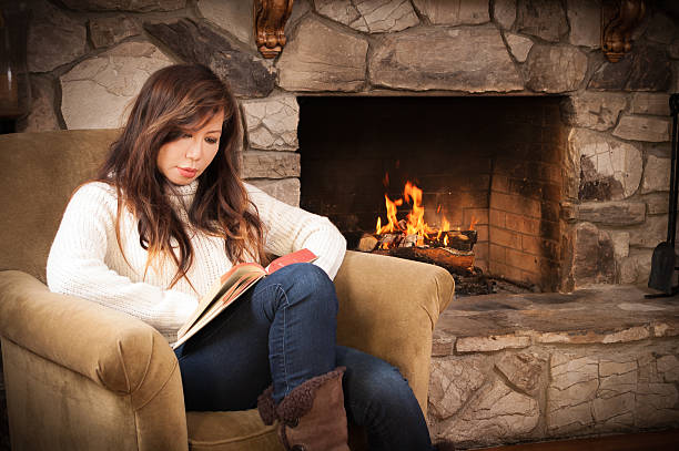 A beautiful Asian woman is sitting next to a roaring fire in a rustic stone fireplace. She is dressed warmly in a white sweater and is reading a book. She's either in her living room or on vacation in a cozy cabin.