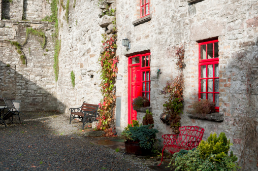 Outdoor of the Abbey Dodge in the Sligo Abbey in the Republic of Ireland. The red door and windows stand out the grey stone wall of the dodge, together with the green leaves of the ivy which decorates the facade. Two benches stand ready to welcome visitors.