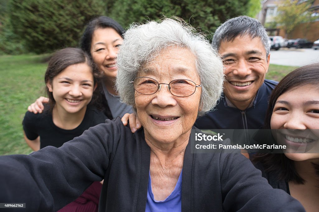 Grandmother, Children, Grandchildren Pose for Selfie, Care Home in Background A fit ,senior, Asian Grandmother smiles with her children and mixed-ethnic grandchildren while posing for a selfie in a park garden.   Real, three generation family including a senior woman, mature adult son and daughter and teenage grand-daughters.  An assisted living facility can be seen in the background.  Camera point of view. Asian and Indian Ethnicities Stock Photo