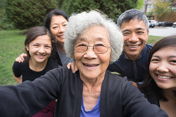 Grandmother, Children, Grandchildren Pose for Selfie, Care Home in Background A fit ,senior, Asian Grandmother smiles with her children and mixed-ethnic grandchildren while posing for a selfie in a park garden.   Real, three generation family including a senior woman, mature adult son and daughter and teenage grand-daughters.  An assisted living facility can be seen in the background.  Camera point of view. canada photos stock pictures, royalty-free photos & images