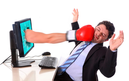 Businessman with computer wearing suit and necktie sitting at office desk hit by boxing glove comming out of laptop monitor in overwork, stress and crisis concept, and also mobbing, bullying, cybermobbing and cyberbullying