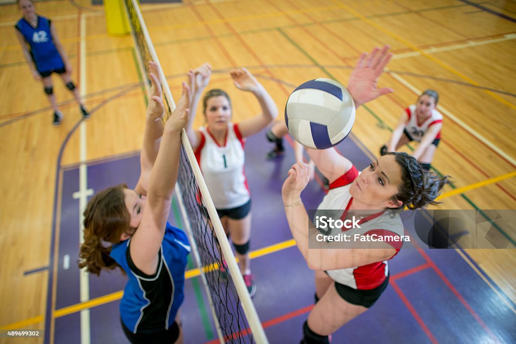 Women Spiking and Blocking a Volleyball A group of teenage women playing in a volleyball match in a school gymnasium. One girl is jumping up to spike the ball. Volleyball - Sport Stock Photo