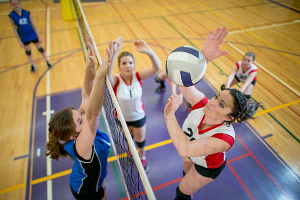 Women Spiking and Blocking a Volleyball A group of teenage women playing in a volleyball match in a school gymnasium. One girl is jumping up to spike the ball. spiked stock pictures, royalty-free photos & images