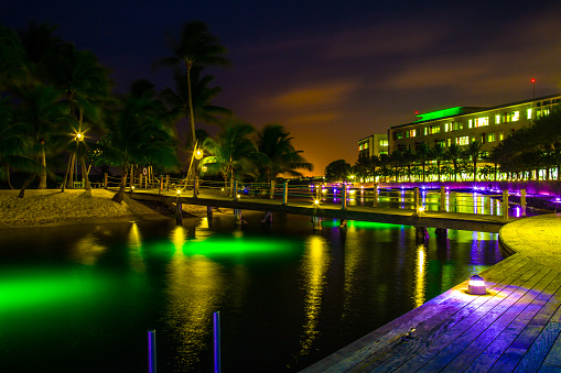 Camana Bay, Cayman Islands - June 18, 2015: It is a beautiful tropical evening  along the waterfront in the town of Camana Bay on Grand Cayman Island. The town of Camana Bay encompasses 600 acres stretching from Seven Mile Beach to North Sound. Camana Bay is a relatively new town that consists of residential neighborhoods, parks, gardens, marinas, restaurants, shops and sports facilities. Colorful lights are lighting up the walkways and footbridges that cross over the water.