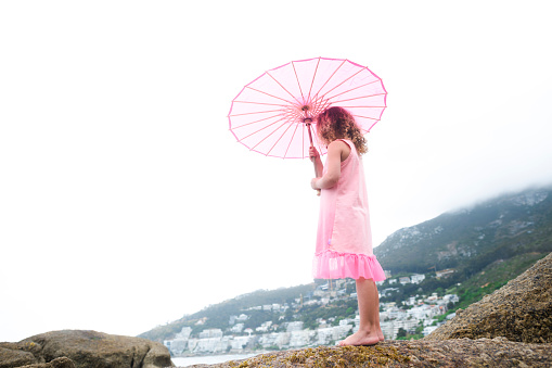 Little girl standing on a boulder at the beach holding a pink parasol.
