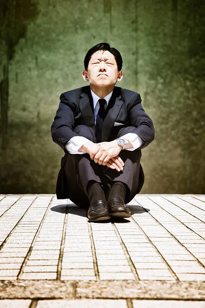 Stressed-out Japanese buisinessman sits on the pavenment and prays, eyes closed.