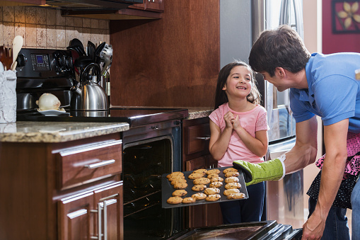 A father in the kitchen with his 6 year old daughter, baking cookies.  The cookies are ready, so he has opened the oven door, and is taking the sheet out with an oven mitt.  They little girl is excited, and they are looking at each other, smiling.