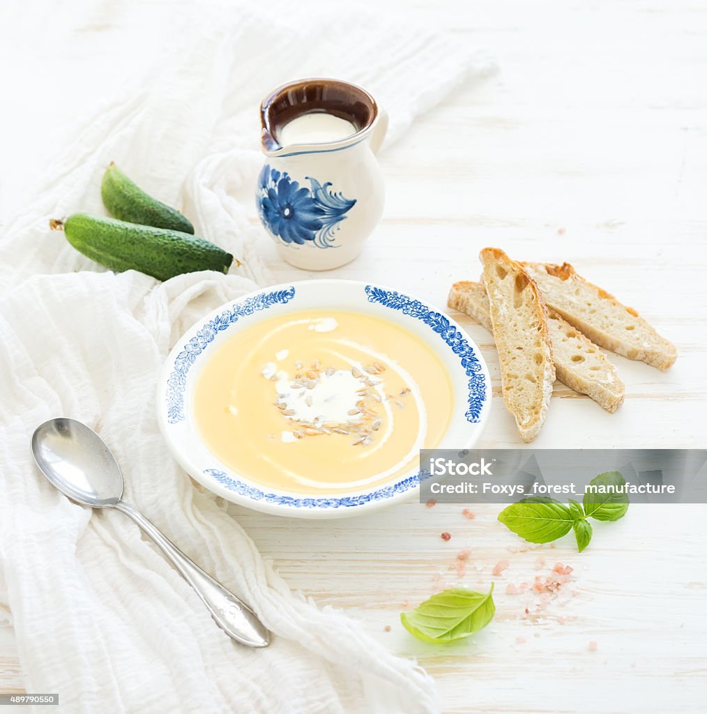 Pumpkin soup with cream, fresh basil, cucumbers and bread in Pumpkin soup with cream, fresh basil, cucumbers and bread in vintage ceramic plate over white wooden background 2015 Stock Photo