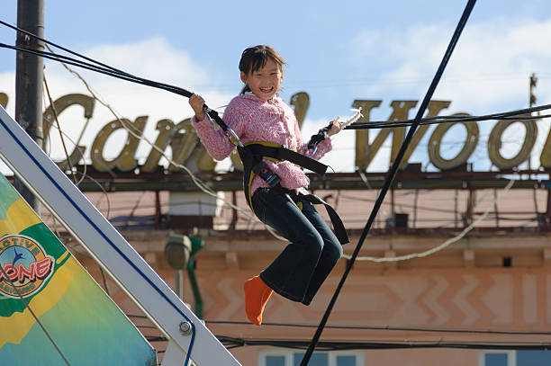 Young girl joyfully performing an acrobatic jump on a bungee trampoline under the bright sun, surrounded by a clear blue sky and fluffy clouds