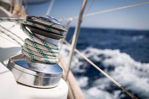 A winch on a sail boat is used to tighten the rope of the front sail. Selective focus on the rope itself, the photograph was taken while the sail boat is under strong wind conditions, with which you can notice the breaking waves and foams in the background.