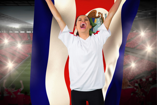 Football fan in white cheering holding costa rica flag against vast football stadium with fans in red