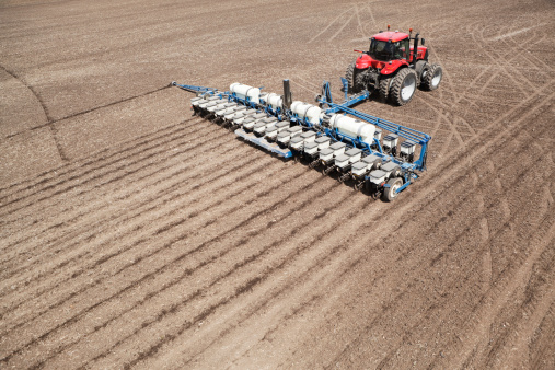 A low aerial view of a tractor planting a Spring cornfield. The 16 row planter has its marker wheel extended for alignment of the next pass. The tanks on the planter hold liquid fertilizer which is applied to the seed row.