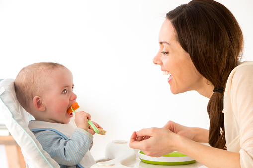 Side view of baby boy in high chair eating using a fork, mum facing him smiling,