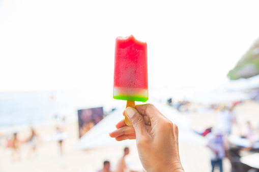 Close up human hands holding an ice cream stick. The ice cream has red color on top and green color below. Its a stick ice cream. The background is a beach in the afternoon.
