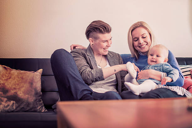 Happy mothers playing with baby on sofa at home Happy mothers playing with baby on sofa. Gay women with cute toddler in living room. Happy family of three at home. unknown gender stock pictures, royalty-free photos & images