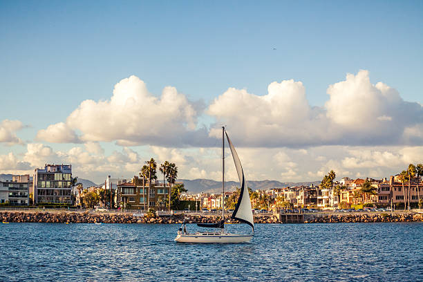 The main channel into Marina Del Rey, with Venice Beach and canal adjacent houses in the background and a sailboat coming in to dock in the foreground.