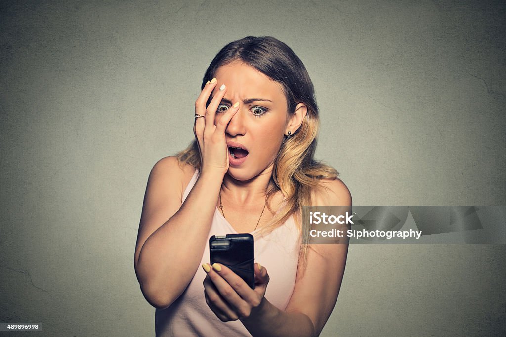 anxious scared girl looking at phone seeing bad news Closeup portrait anxious scared young girl looking at phone seeing bad news photos message with disgusting emotion on her face isolated on gray wall background. Human reaction, expression Telephone Stock Photo