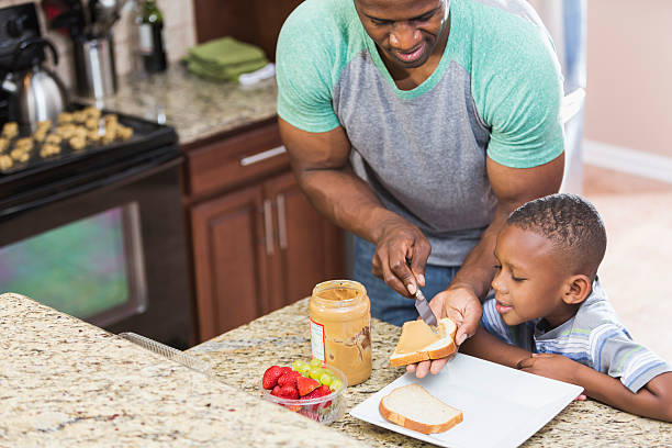 Father at home making peanut butter sandwich for son An African American single father taking care of his 5 year hold son, making a peanut butter sandwich for lunch as the little boy watches eagerly.  They are standing in the kitchen looking down at the slice of bread in the man's hand. role reversal stock pictures, royalty-free photos & images