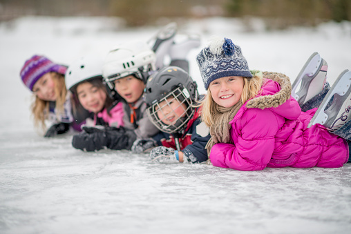 A group of children are lying in a row on the ice wearing hockey helmets. They are smiling and looking at the camera.