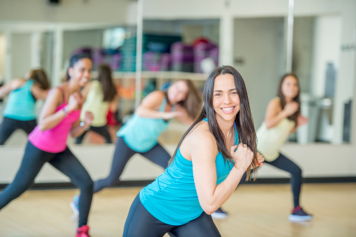 A group of women are dancing together in a fitness class at a gym studio. They are wearing fitness clothing and are smiling while looking at the camera.