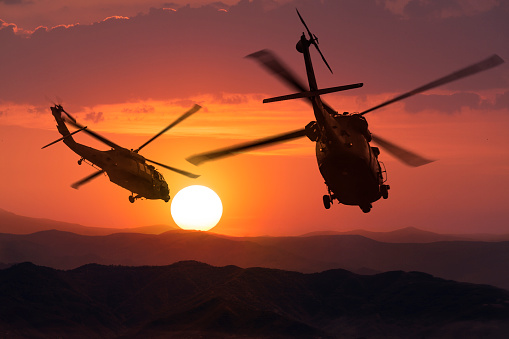 Silhouette aerial view of Army helicopters flying over mountains at sunset. 