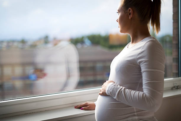 bien femme enceinte heureuse, regardant par la fenêtre - être enceinte photos et images de collection