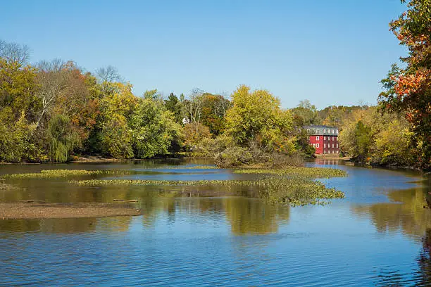 Fall foliage is reflected in the water at Delaware and Raritan Canal State Park in Princeton, New Jersey.  An old red mill house can be seen in the background.