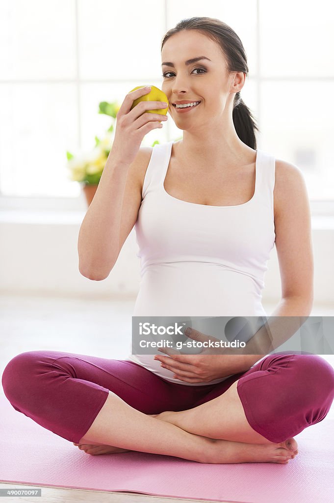 Only healthy eating for both of them. Beautiful pregnant woman sitting in lotus position and eating apple Adult Stock Photo