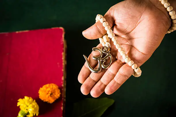 open hand showing Aum locket with beads, red hindu book in the background with flowers