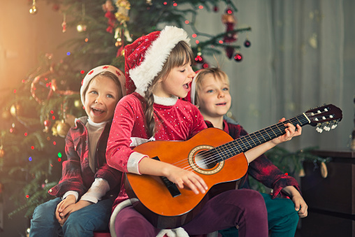 Little boys and little girl wearing santa's hat singing carols near christmas tree. The boys are aged 5 and the girl is aged 8. The girls is playing a guitar.