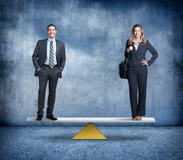 Gender Equality In The Workplace A businessman and businesswoman stand on opposite ends of a perfectly balanced seesaw.  Each is standing and smiling at the camera. gender equality stock pictures, royalty-free photos & images