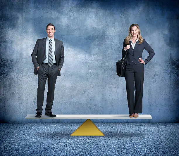 A businessman and businesswoman stand on opposite ends of a perfectly balanced seesaw.  Each is standing and smiling at the camera.