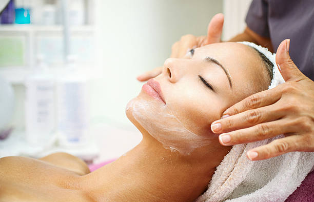 Facial treatment at beauty salon. Closeup of adult caucasian woman having her face pampered at beauty salon by unrecognizable mid aged professional female beautician. The client is relaxing on massage bed wrapped in white towel. Some cosmetic products are on the shelves in background, blurry. facial massage stock pictures, royalty-free photos & images